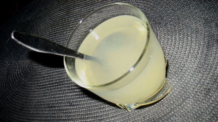 Ein Glas Zitronenwasser steht auf einem dunklen Tisch und reflektiert das Licht, w&auml;hrend ein L&ouml;ffel darin liegt.