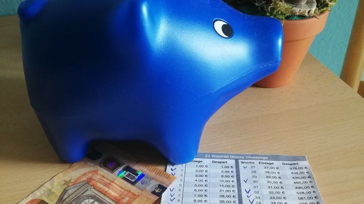 Ein blauer Sparschwein steht auf einem Tisch, neben ihm liegt ein 50-Euro-Schein und eine Liste mit Sparbetr&auml;gen.