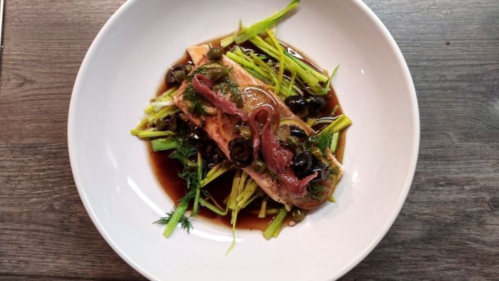 Zarte Lachsfilets liegen auf einem Bett aus frischem Gem&uuml;se in einer eleganten wei&szlig;en Sch&uuml;ssel, serviert auf einem Holztisch.