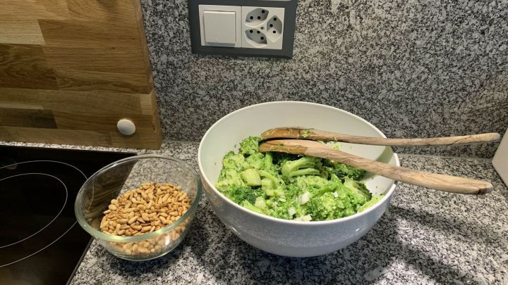 Ein frischer Brokkolisalat liegt in einer gro&szlig;en wei&szlig;en Sch&uuml;ssel auf einer grauen Arbeitsplatte, daneben eine Schale mit ger&ouml;steten N&uuml;ssen.