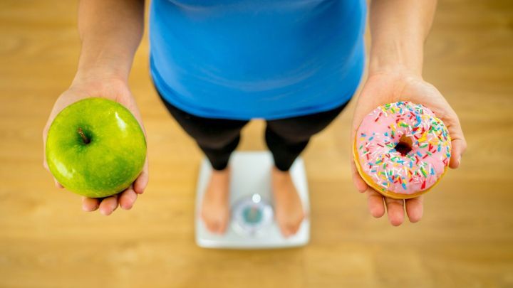 Eine Person steht auf einer Waage und h&auml;lt einen gr&uuml;nen Apfel in der linken Hand und einen bunten Donut in der rechten, was die Entscheidung zwischen gesunder Ern&auml;hrung und Leckereien symbolisiert.
