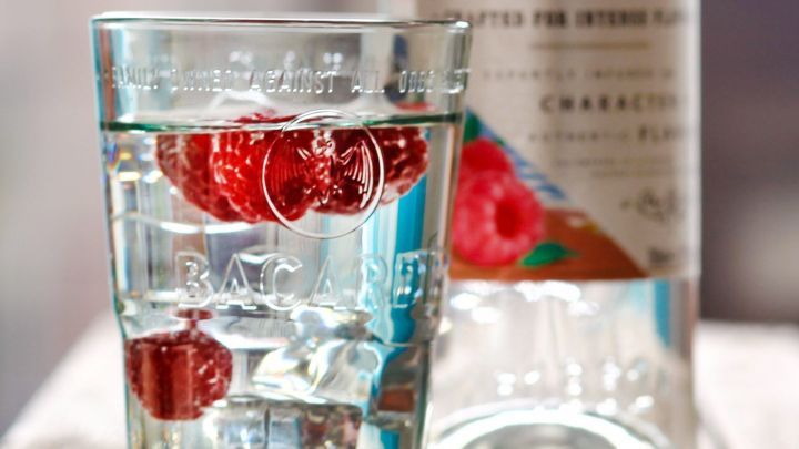 Ein Glas mit Bacardi Razz und frischen Himbeeren steht auf einem Holztisch, umgeben von einem modernen Raum.