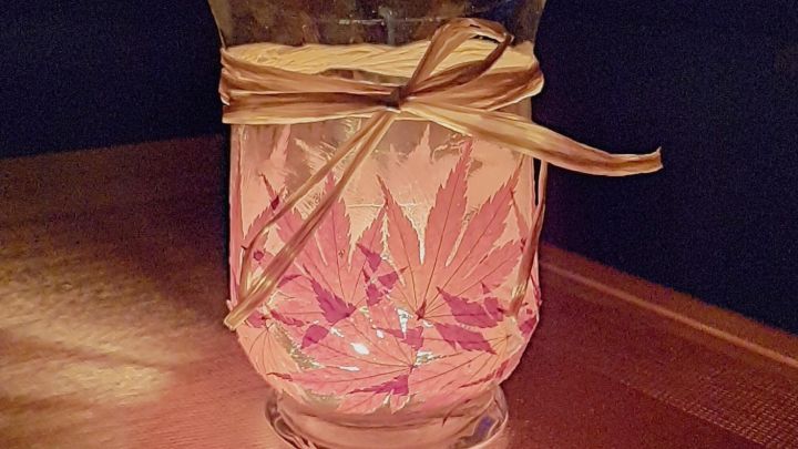 Ein dekoratives Windlicht mit Bl&auml;ttern und einem Band steht auf einem Tisch, umgeben von d&auml;mmrigem Licht im Raum.