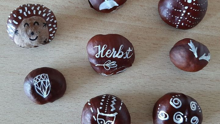 Kastanien sind bunt bemalt und liegen kreativ auf einem Tisch, darunter sind verschiedene Muster und das Wort "Herbst".