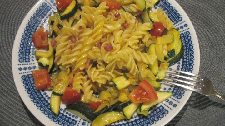 Bunte Schinken-Nudeln liegen auf einem Teller, begleitet von frischem Gem&uuml;se in einer hellen, gem&uuml;tlichen K&uuml;che.