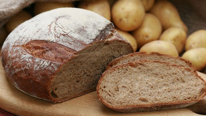Ein frisch gebackenes Kartoffel-Hausbrot liegt samt einer Scheibe auf einem Holzbrett, umgeben von frischen Kartoffeln.