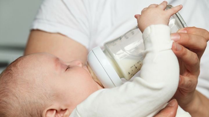 Ein Baby h&auml;lt eine Flasche mit Milch und trinkt daraus, w&auml;hrend es von einer Person in einer gem&uuml;tlichen Umgebung gehalten wird.