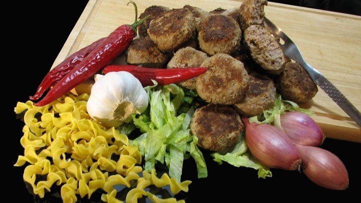 Vielzahl von Frikadellen mit Linsen liegt auf einem Holzbrett, umgeben von roten Chilis, Knoblauch und bunten Nudeln.