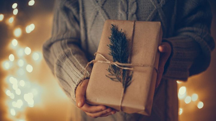 Ein sorgf&auml;ltig verpacktes Geschenk mit Naturmaterialien wird von einer Person in einem warmen, weihnachtlichen Umfeld gehalten.