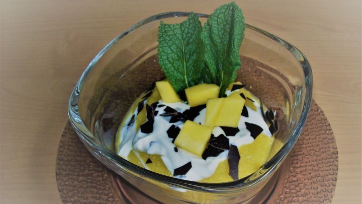 Ein durchsichtiger Becher mit Mango-Quark-Dessert, garniert mit frischen Mangost&uuml;cken und Minzbl&auml;ttern.