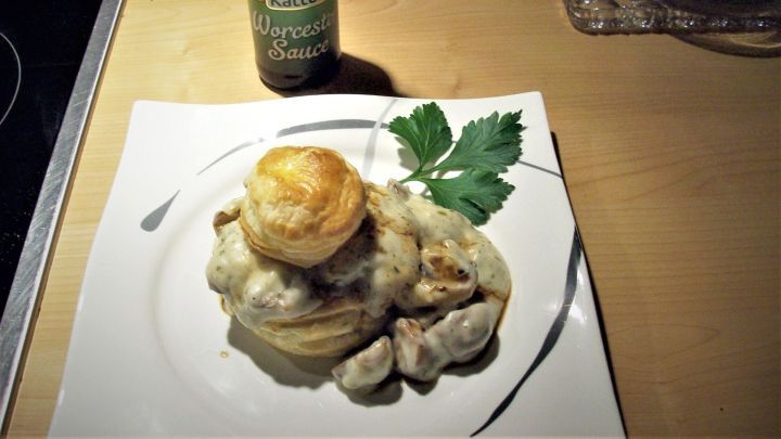 Auf einem wei&szlig;en Teller liegen goldbraune Champignon-Pasteten, umgeben von einer cremigen Sauce und frischer Petersilie. Daneben steht ein Gew&uuml;rzglas.