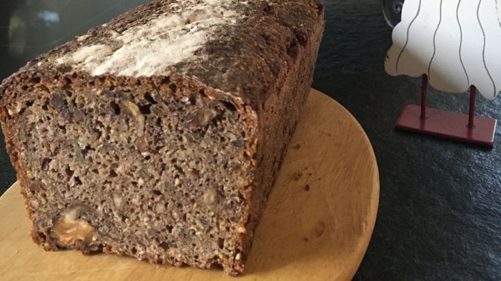 Ein Nussbrot liegt auf einem Holzbrett und wird von einer Obstschale mit &Auml;pfeln und Orangen umgeben.