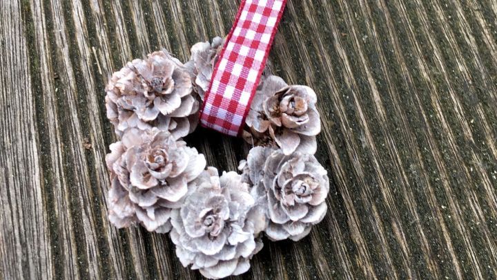 Ein Minikranz aus Tannenzapfen h&auml;ngt an einem karierten Band und liegt auf einer Holzoberfl&auml;che.