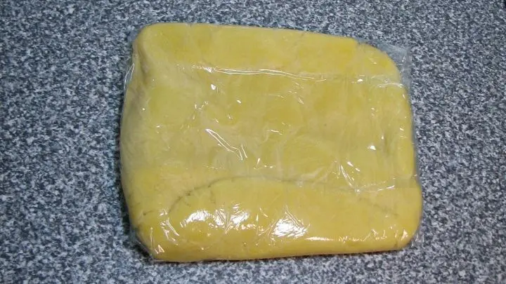 Aus den Kr&uuml;meln mit den H&auml;nden einen geschmeidigen Teig zusammen kneten, auf Frischhaltefolie legen, einer flachen Platte dr&uuml;cken. Rechteckig formen und darin einschlagen.