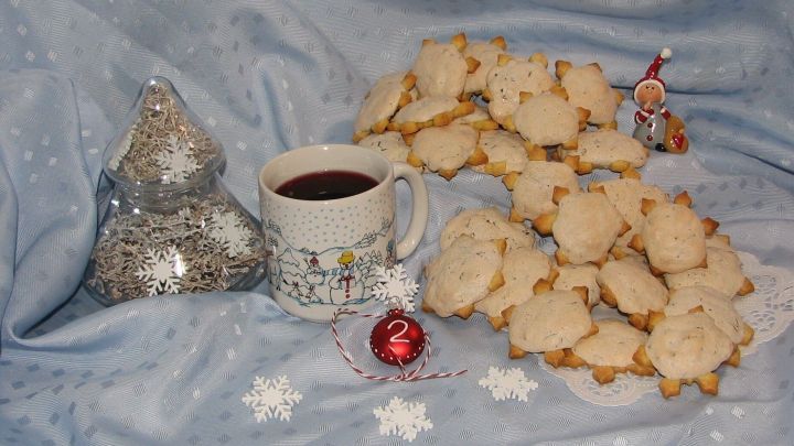 Auf einem blauen Tisch liegen zahlreiche Schneekristalle-Kekse, neben einer Tasse Gl&ouml;gg und festlichem Schmuck.