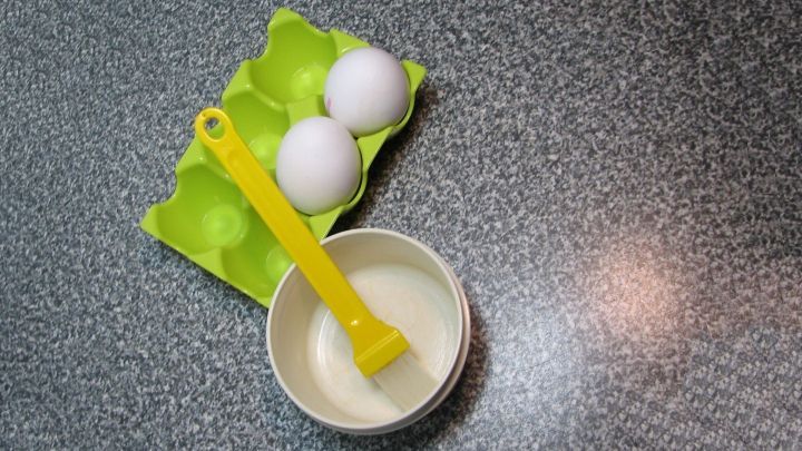 Zwei frische Eier liegen in einem gr&uuml;nen Eierkarton, w&auml;hrend eine gelbe Beschichtungsspritze in einer wei&szlig;en Sch&uuml;ssel mit Eiwei&szlig; steht.