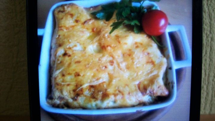 Eine goldbraune Portion gratinierte Spinat-Lachs-Pfanne liegt in einer wei&szlig;en Auflaufform auf einem Holzbrett, garniert mit Petersilie und einer Kirschtomate.