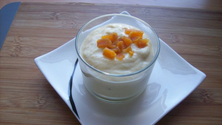 Ein Glas Mango-Joghurt mit Aprikosenst&uuml;cken steht auf einem wei&szlig;en Teller, umgeben von einer Holzplatte.