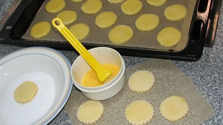 Zucker in eine flache Schale geben. Für die Eistreiche das Eigelb mit der Sahne etwas Zucker (½ TL) und einer Prise Salz verkleppern. Die Taler damit dünn einstreichen und kopfüber in den Zucker drücken.