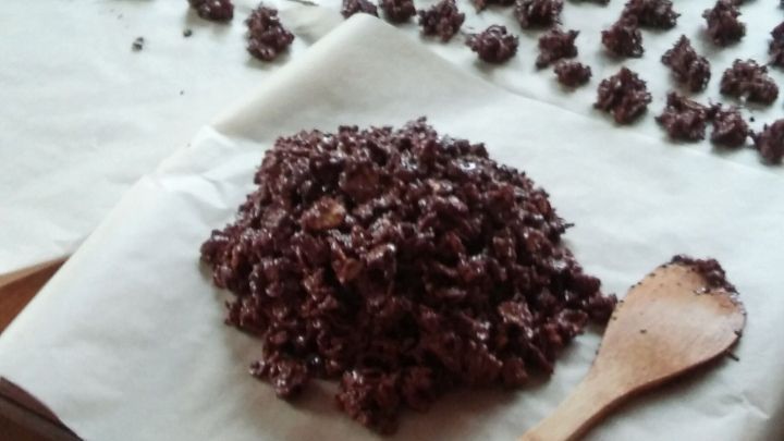 Eine Vielzahl von Schoko-Crossies liegt auf Backpapier, w&auml;hrend eine Holzspatel neben einer gro&szlig;en Menge Schokoladenmasse steht.