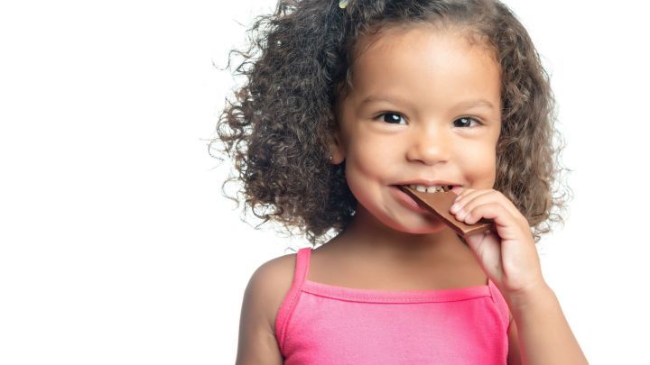 Ein l&auml;chelndes Kind mit lockigem Haar bei&szlig;t in ein St&uuml;ck Schokolade, was Freude und Genuss vermittelt.