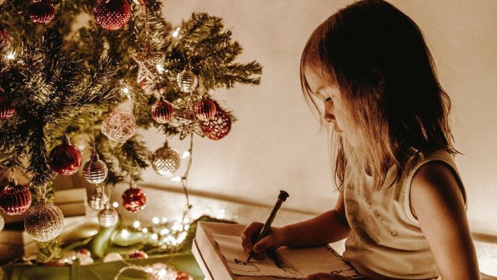 Ein M&auml;dchen sitzt umgeben von Lichtern und einem Weihnachtsbaum, w&auml;hrend es in ein Notizbuch schreibt.