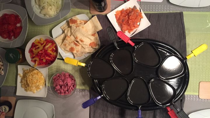Ein Raclette-Grill liegt auf einem Tisch, umgeben von vorbereiteten Zutaten wie Gem&uuml;se, K&auml;se und Saucen f&uuml;r Pizza-Raclette.