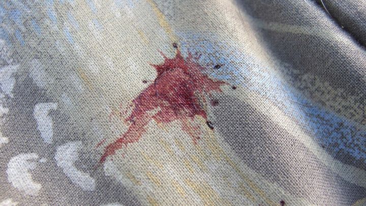 Ein Blutfleck breitet sich auf einem grauen Stoff aus, umgeben von gemusterten Verzierungen.