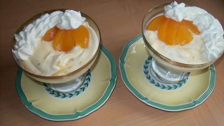 Zwei Schalen mit Aprikosen-Sahne-Schaumspeise, garniert mit Sahne und einer Aprikose, stehen auf einem Tisch.
