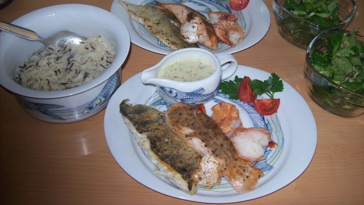 Auf den Tellern wird zart gegrillter Fisch mit Wildreis und frischem Salat serviert, umgeben von feinen Saucen und Kr&auml;utern.