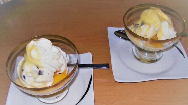 Zwei Dessertschalen mit Stracciatella-Eis und gelber Fruchtsauce stehen auf einer Tischplatte aus Holz.