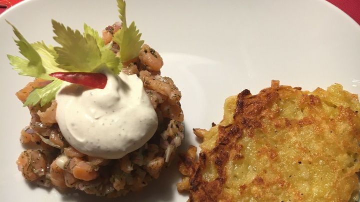 Ein Lachstatar wird auf einem wei&szlig;en Teller serviert, garniert mit Sauerrahm und frischen Kr&auml;utern, daneben zwei knusprige R&ouml;stis.