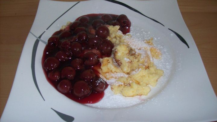 Ein Teller zeigt Grie&szlig;schmarrn, der neben einer gro&szlig;z&uuml;gigen Portion Kirschkompott angerichtet ist, garniert mit Puderzucker.