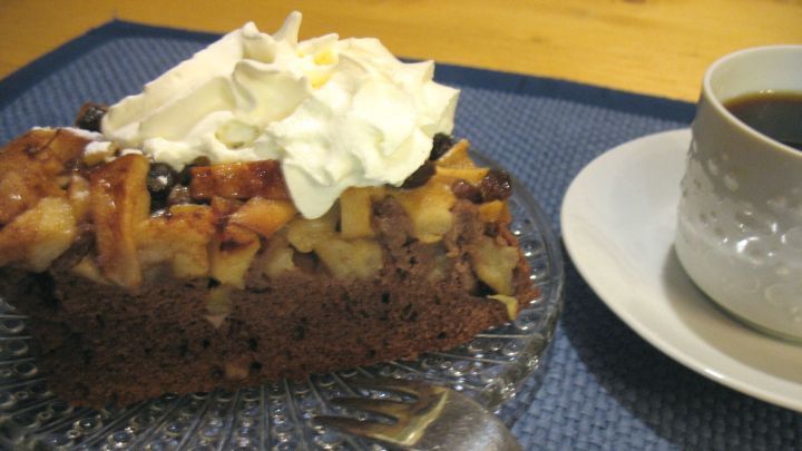 Saftiger Apfelkuchen mit einer dunklen Bodenlage liegt auf einem Glasteller neben einer Tasse Kaffee, dekoriert mit Sahne.