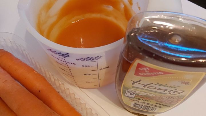 Eine Mischung aus Karottenbrei und Honig steht in einem Becher auf einem Tisch, um Hustenreiz zu lindern. Karotten liegen daneben.