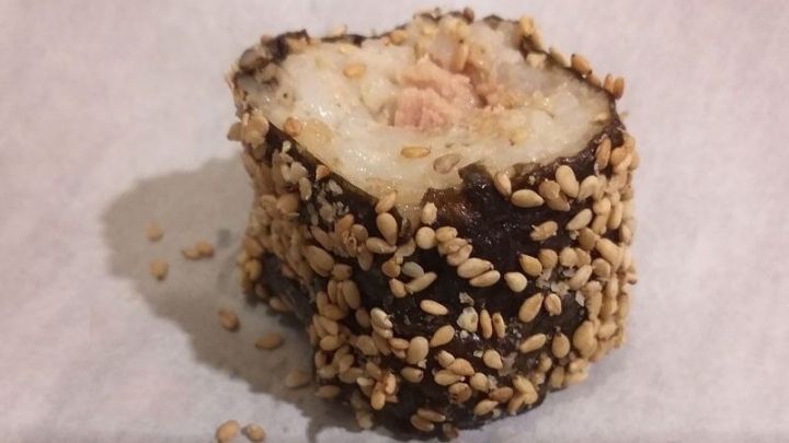 Ein St&uuml;ck Sushi liegt auf einem wei&szlig;en Papier. Es besteht aus Reis und ist mit Sesam umh&uuml;llt, wirkt frisch zubereitet.