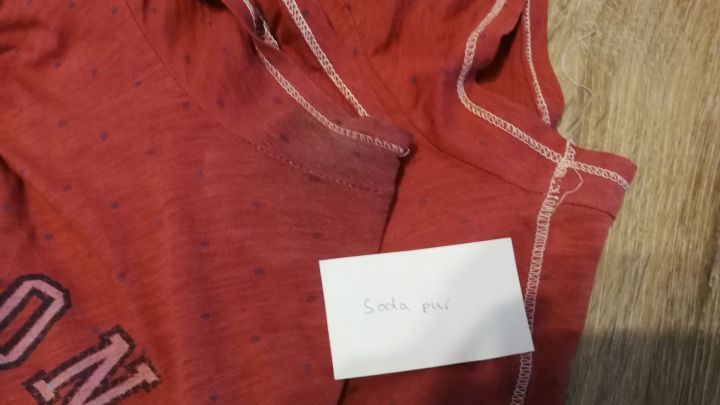 Ein rotes T-Shirt mit Deoflecken liegt auf einem hellen Untergrund, daneben befindet sich ein kleiner Notizzettel.