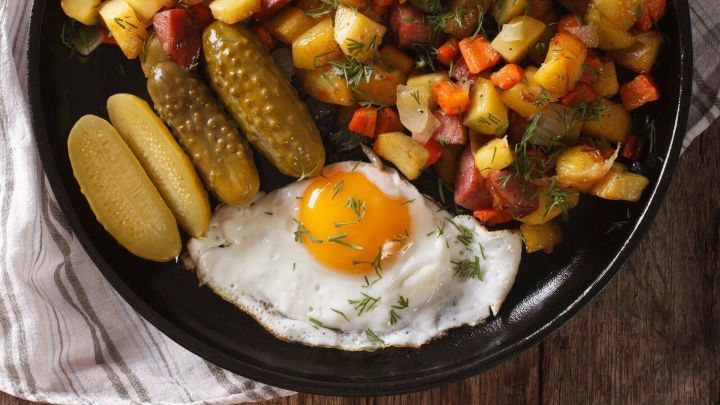 Ein schwarzer Teller mit einer bunten Mischung aus Gem&uuml;se, Kartoffeln, Speck und einem Spiegelei liegt auf einem Holzuntergrund.