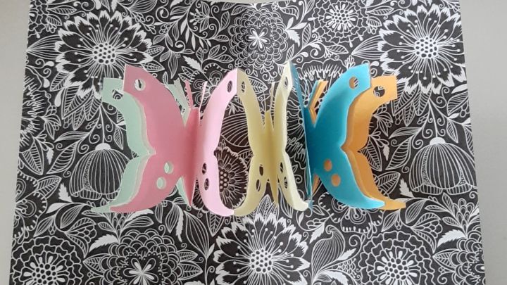 Bunte Schmetterlinge stehen in einer Pop-Up-Karte, die auf einem floralen Hintergrund aufgeklappt ist.