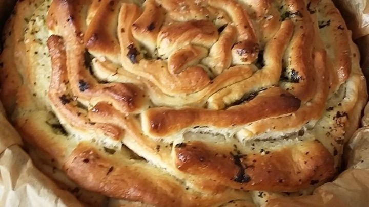 Ein frisch gebackenes Zupfbrot mit F&uuml;llung liegt in einer runden Form. Es hat eine sch&ouml;ne spiralf&ouml;rmige Oberfl&auml;che und befindet sich auf einem wei&szlig;en Tisch.