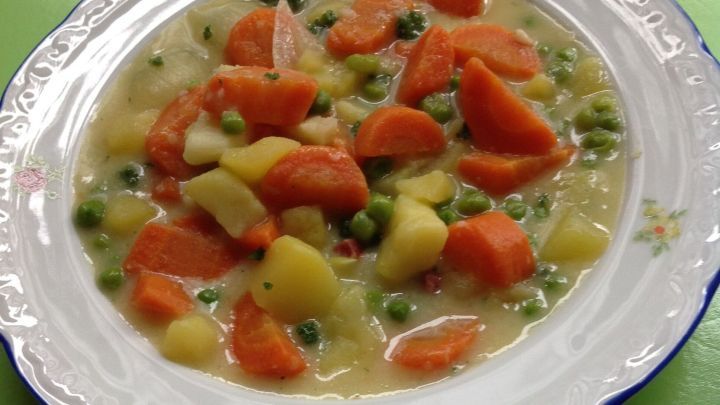 Ein Teller mit feiner Gem&uuml;sesuppe liegt auf einer gr&uuml;nen Unterlage, gef&uuml;llt mit Karotten, Erbsen und Kartoffeln.