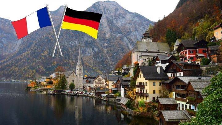 Zwei Flaggen, eine franz&ouml;sische und eine deutsche, wehen vor einer malerischen Stadt am See, umgeben von Bergen.