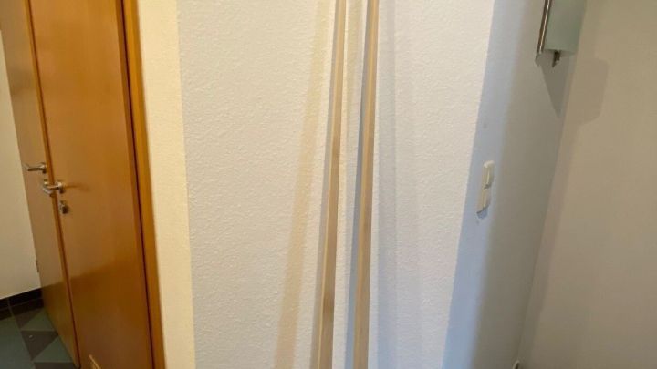 Drei lange Holzlatten stehen in einer Ecke eines Flurs neben einer geschlossenen T&uuml;r und grauen Fliesen.