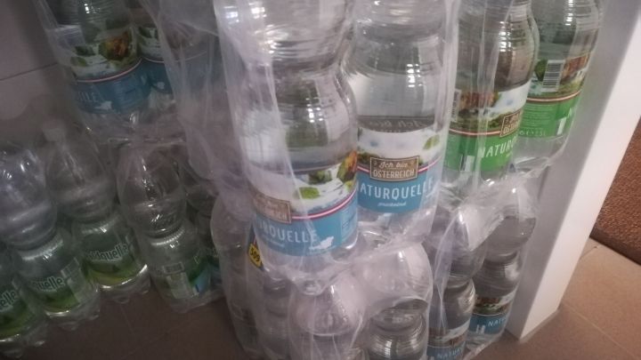 Vor einem Regal sind gestapelte Wasserflaschen zu sehen, die zur Konservierung von Trinkwasser f&uuml;r Notf&auml;lle bereitstehen.