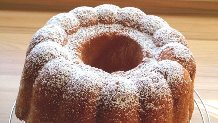 Ein Quark-Gugelhupf aus Hefeteig steht auf einem K&uuml;hlgitter, bedeckt mit Puderzucker vor einem hellen Fenster.