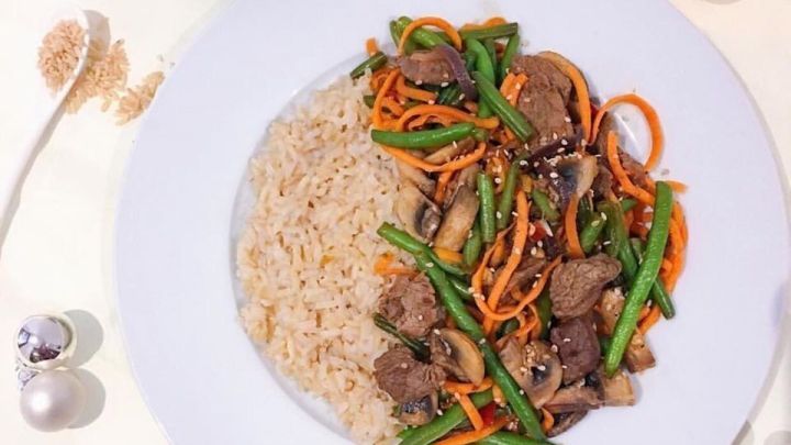 Eine bunte Asia-Rindfleisch-Pfanne mit Gem&uuml;se liegt auf einem wei&szlig;en Teller, umgeben von Beilagen und Dekoration.