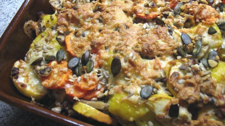 Ein goldbraun gebackener Kartoffel-Apfel-Auflauf mit Katenschinken, garniert mit K&uuml;rbiskernen, pr&auml;sentiert sich appetitlich in einer Auflaufform.