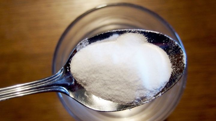 Ein L&ouml;ffel mit wei&szlig;em Natron schwebt &uuml;ber einem Glas, w&auml;hrend es in einer Holzumgebung f&uuml;r Zahnpflege genutzt wird.