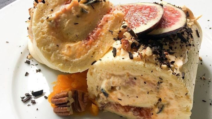 Eine K&uuml;rbis-Biskuitrolle liegt auf einem wei&szlig;en Teller, dekoriert mit Feigen und gehackten N&uuml;ssen, umgeben von Br&ouml;seln.