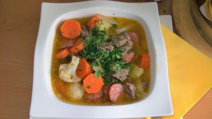 Ein bunter Gem&uuml;setopf mit Karotten, Kartoffeln und Fleisch liegt in einer wei&szlig;en Sch&uuml;ssel auf einem Tisch, umgeben von Besteck.